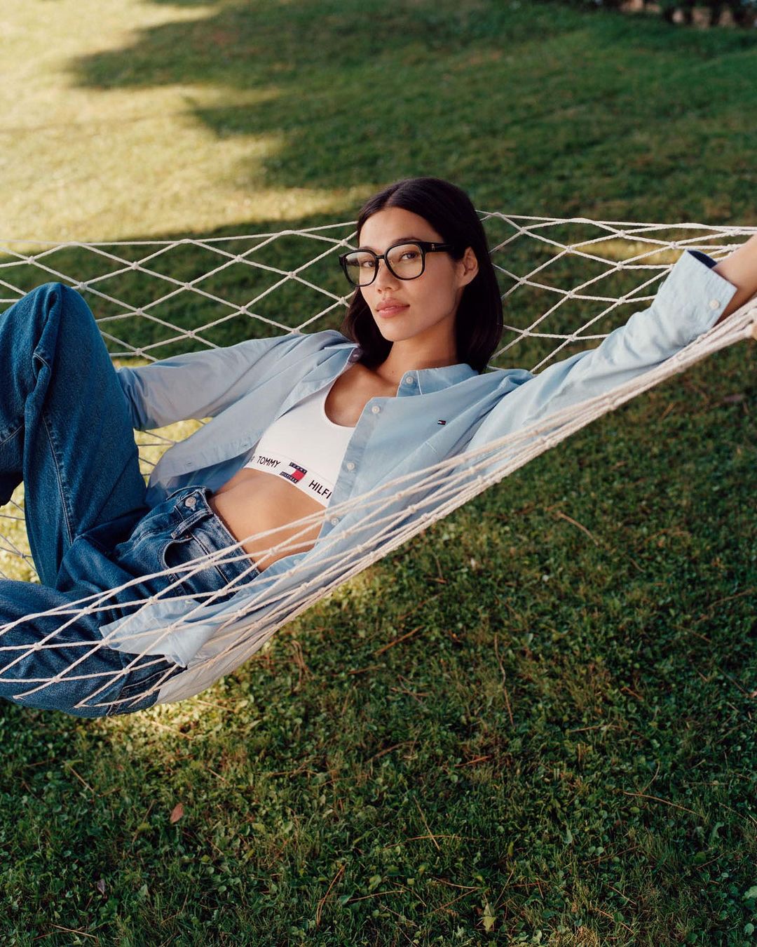A woman lounges in a hammock outside on a lawn, wearing glasses, a white Tommy Hilfiger crop top, and a light blue button-down shirt.