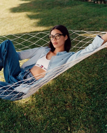 A woman lounges in a hammock outside on a lawn, wearing glasses, a white Tommy Hilfiger crop top, and a light blue button-down shirt.
