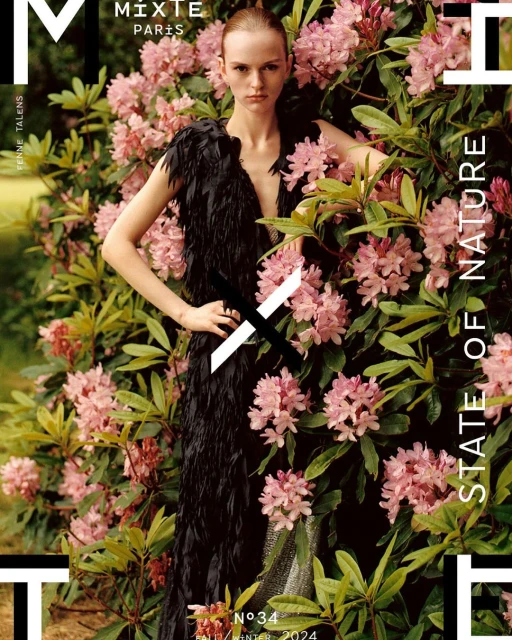 Fashion editorial shot of a fair-skinned adult woman posing outdoors surrounded by blooming pink rhododendron bushes.