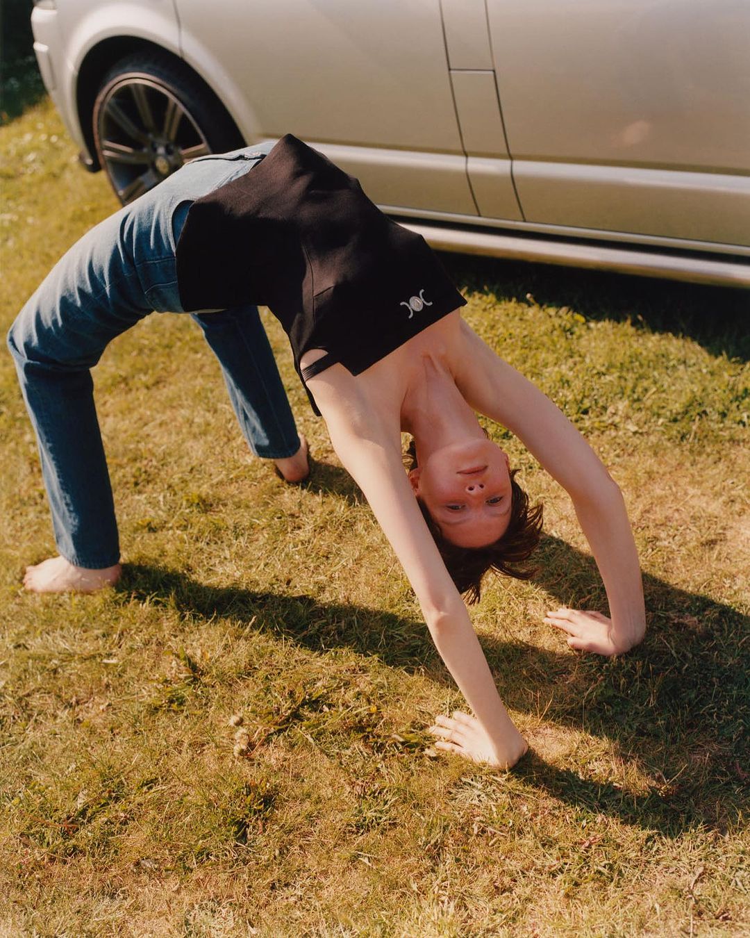 Adult female in a backbend on grass near a silver van, wearing a black sleeveless top and blue jeans, showcasing flexibility outdoors.
