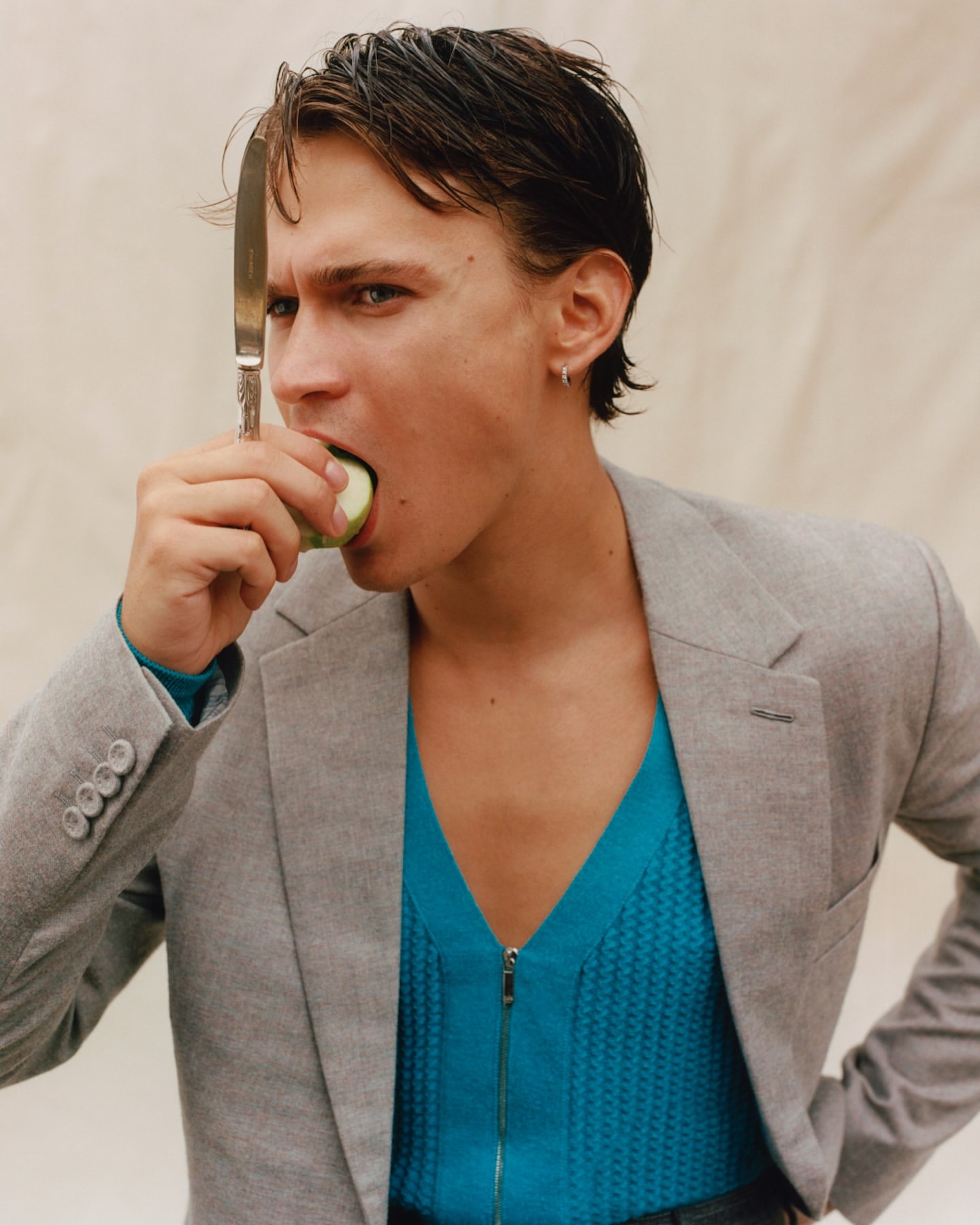 Stylish man bites into a green apple holding a knife, wearing a grey blazer and a teal zippered shirt, captured in a studio setting.