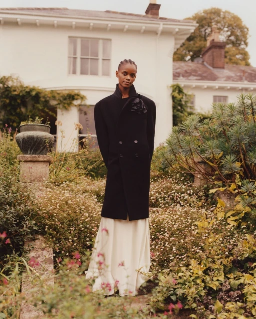 Elegant woman in long black coat and white dress posing in garden with white house backdrop, captured in soft, natural light.