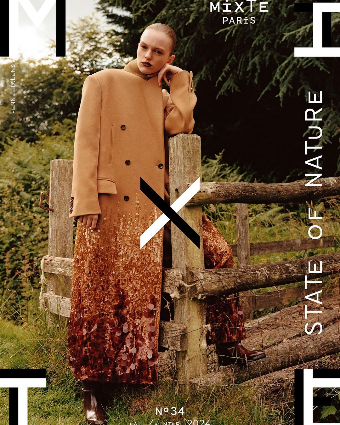 Fashion editorial shot: Woman in a tan coat with decorative bottom stands near a wooden fence in an outdoor, natural setting.