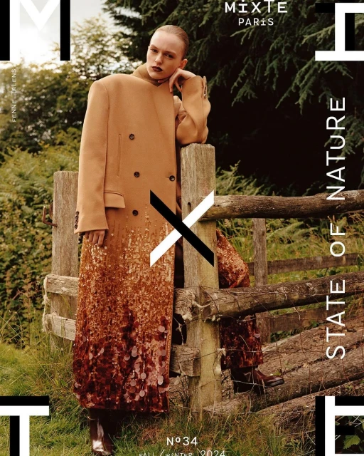 Fashion editorial shot: Woman in a tan coat with decorative bottom stands near a wooden fence in an outdoor, natural setting.