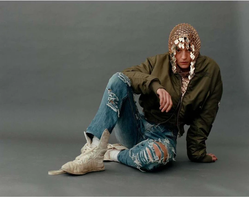 Woman in olive bomber jacket sits with ripped jeans and gold sequin hood. Grey studio backdrop. Fashion editorial shot.