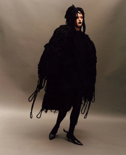 Androgynous fashion model in black, posing in a black fringed robe and black pointed shoes in a studio setting.