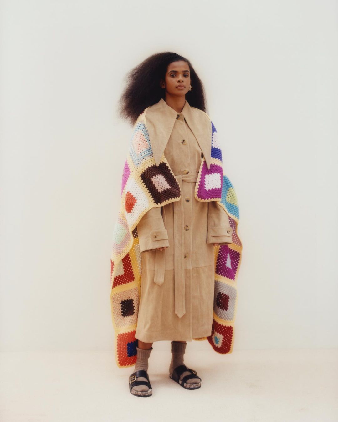 Fashionable woman in trench coat and colorful crochet blanket, posing in a minimalist studio, showcasing unique style and texture contrast.