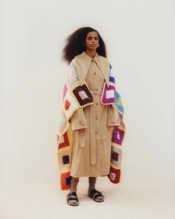 Model wearing a tan coat and a colourful crocheted blanket draped over shoulders, standing against a neutral backdrop