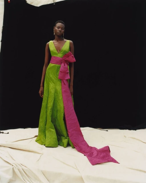 A Black female model poses in a green V-neck gown with a fuchsia sash and bow against a dark backdrop in a studio setting.