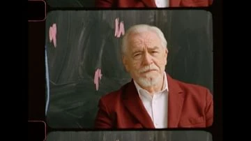 Portrait of an elderly Caucasian man with white hair and beard, wearing a red blazer against a blurry dark background with pink markings.