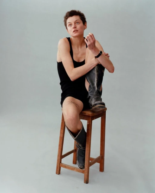 Young adult woman with short hair sits on a wooden stool wearing a black dress and black boots in a neutral-toned studio setting.