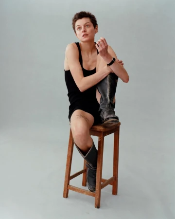 Young adult woman with short hair sits on a wooden stool wearing a black dress and black boots in a neutral-toned studio setting.