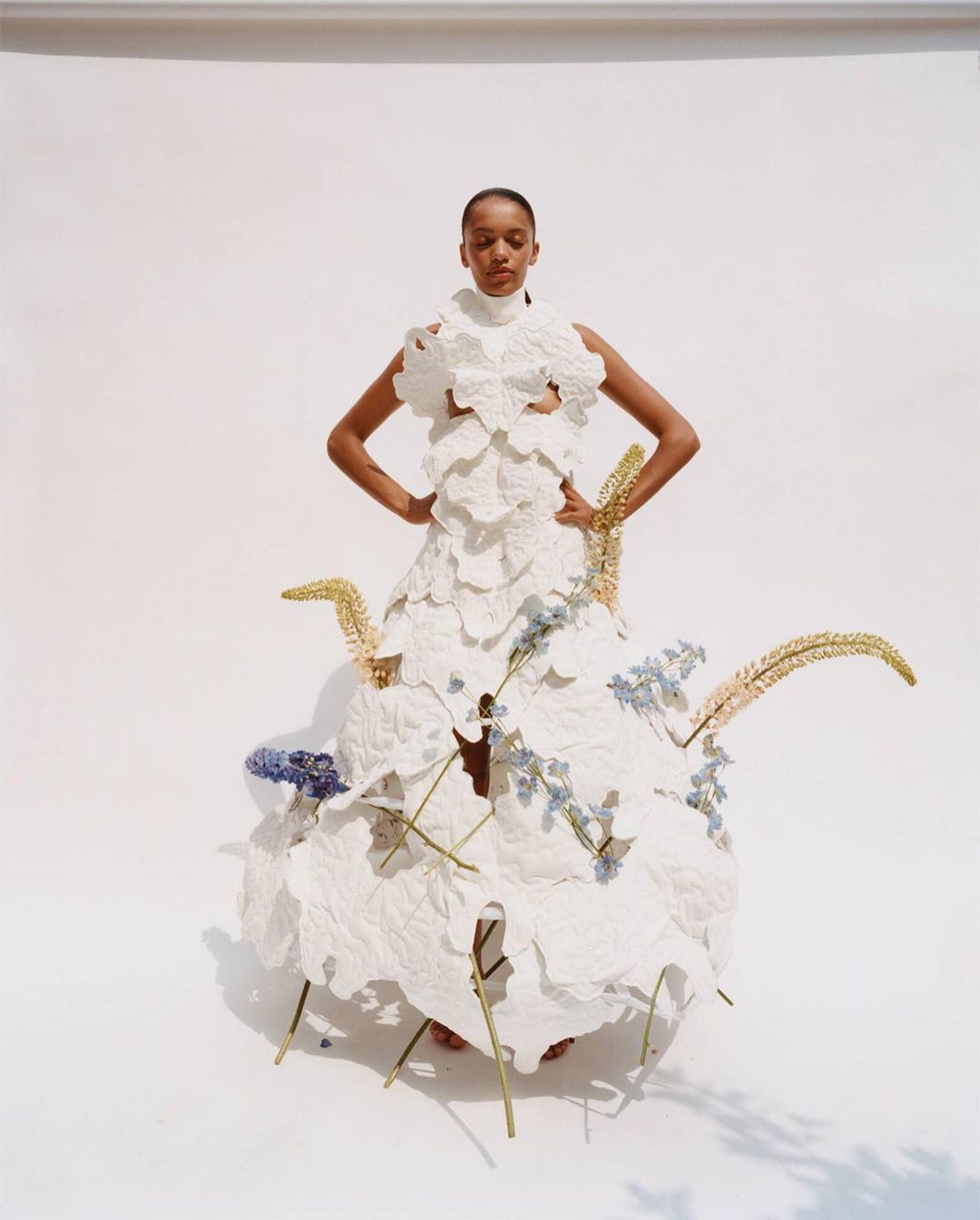 Fashion model poses in a statement dress made from white fabric and floral details in a brightly lit photo studio against a white backdrop.