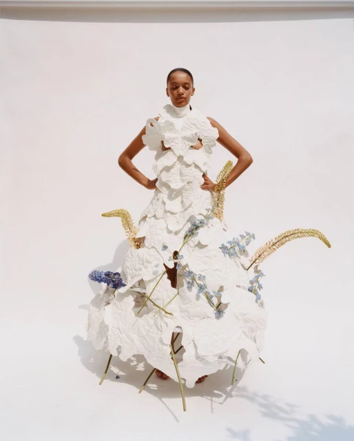 Fashion model poses in a statement dress made from white fabric and floral details in a brightly lit photo studio against a white backdrop.