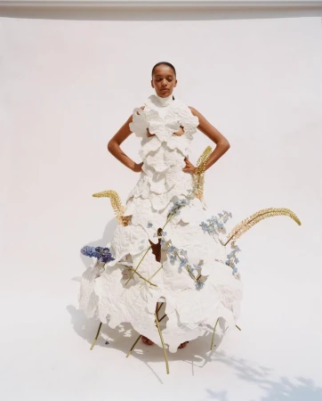 Fashion model poses in a statement dress made from white fabric and floral details in a brightly lit photo studio against a white backdrop.