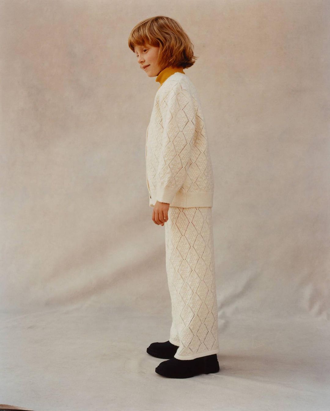A child model with blonde hair poses in a long-sleeved white cardigan and pants with a diamond pattern against a neutral backdrop.