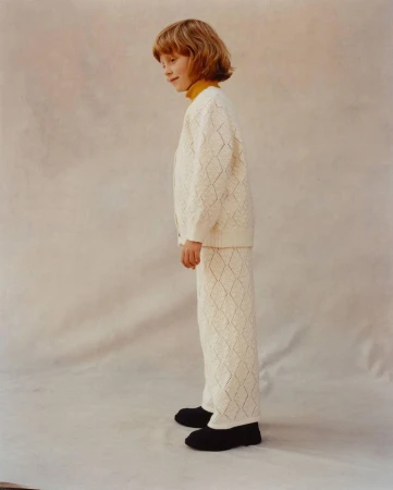 A child model with blonde hair poses in a long-sleeved white cardigan and pants with a diamond pattern against a neutral backdrop.