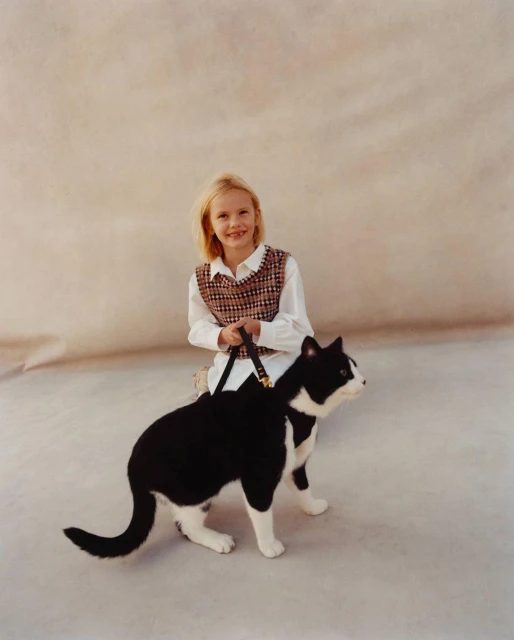 A young girl kneels in a photo studio, holding the leash of a tuxedo cat with white paws. The background is mottled neutral tones.