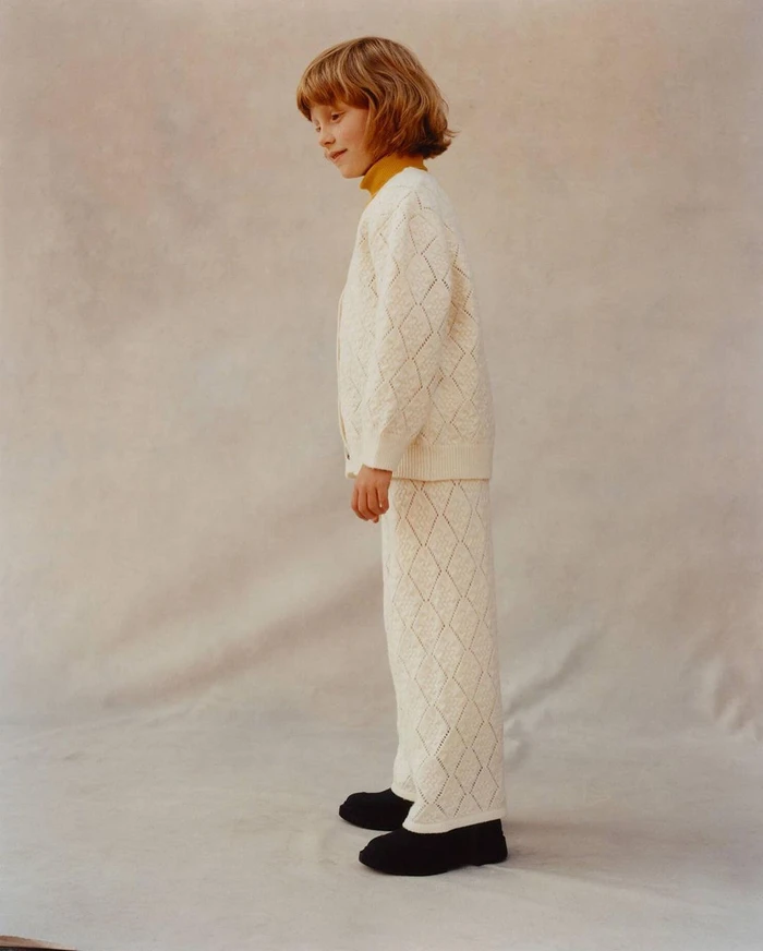Young child in cream knit patterned cardigan and matching trousers standing against a textured neutral background