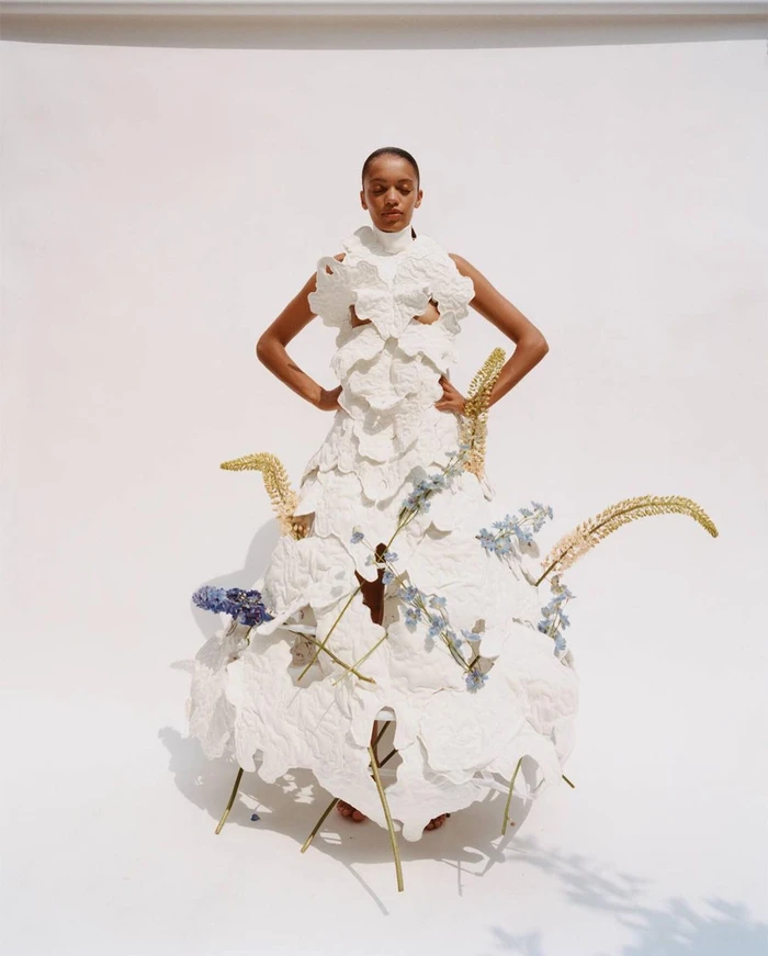 Model wearing a white sculptural textured dress adorned with flowers, photographed by Ben Parks against a white background.