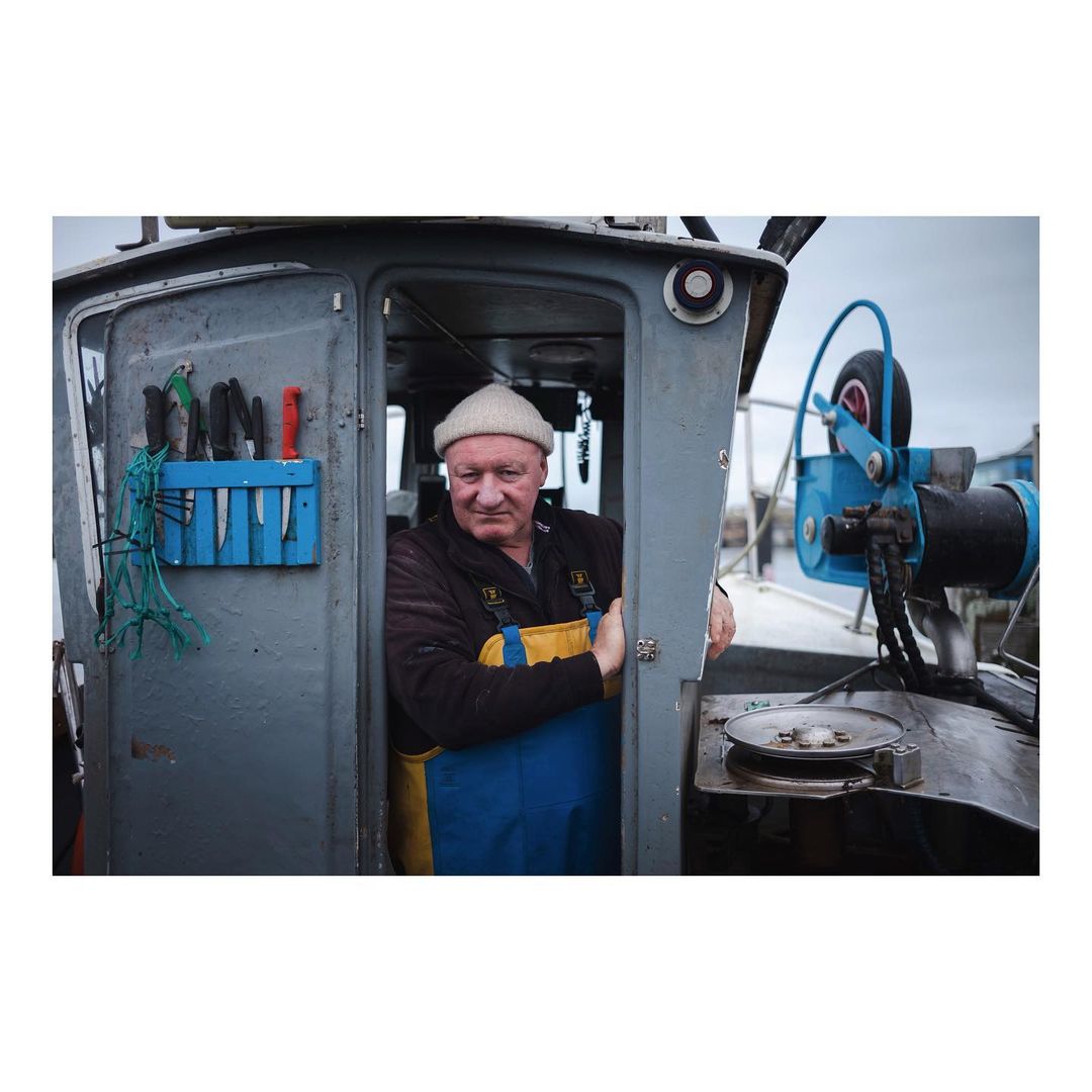 Adult male stands inside the cabin of a fishing boat wearing a knit cap and blue overalls with fishing equipment visible on the boat.