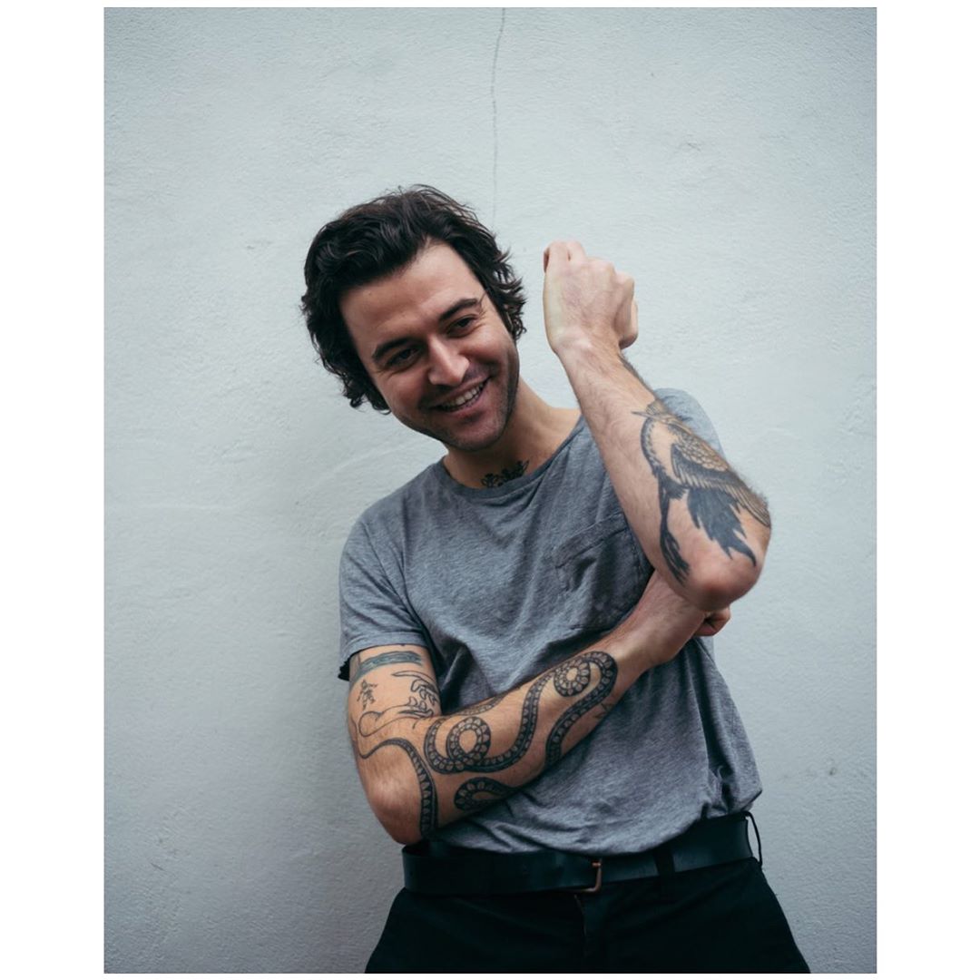 Smiling man with tattoos on his arms poses in front of a white wall, wearing a grey t-shirt and black pants with a dark belt.