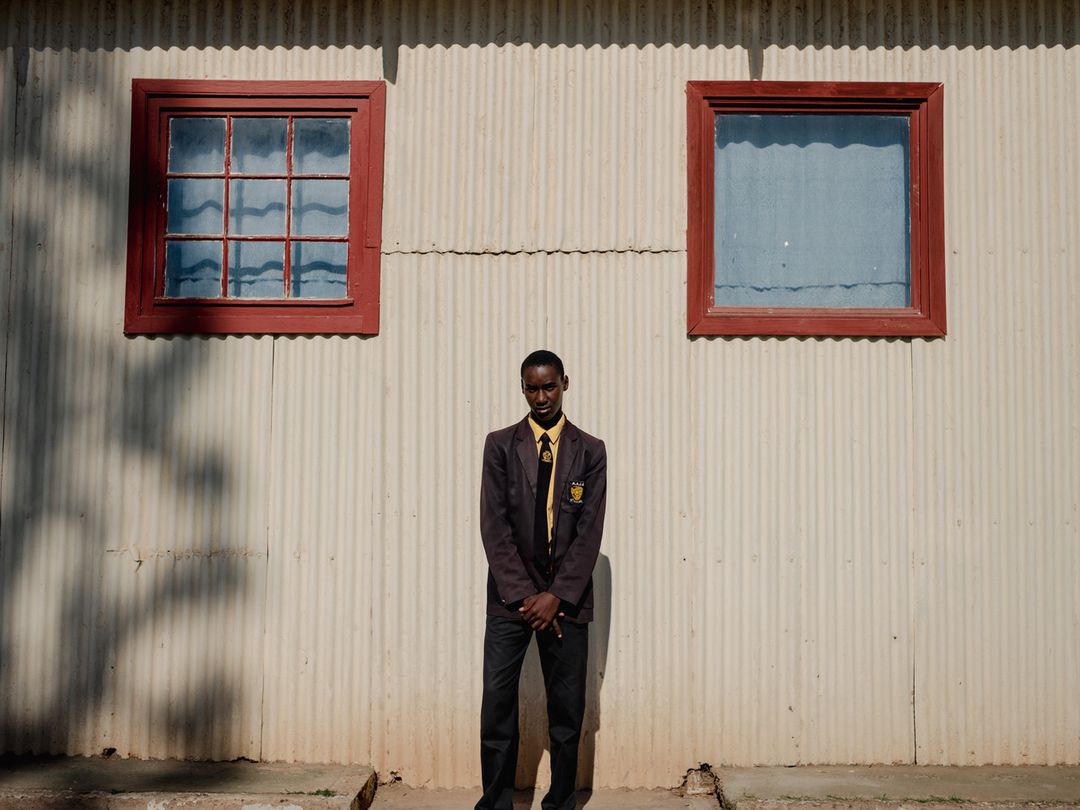 Young adult stands in uniform against textured wall with symmetrical windows casting shadow. The scene features symmetrical composition.