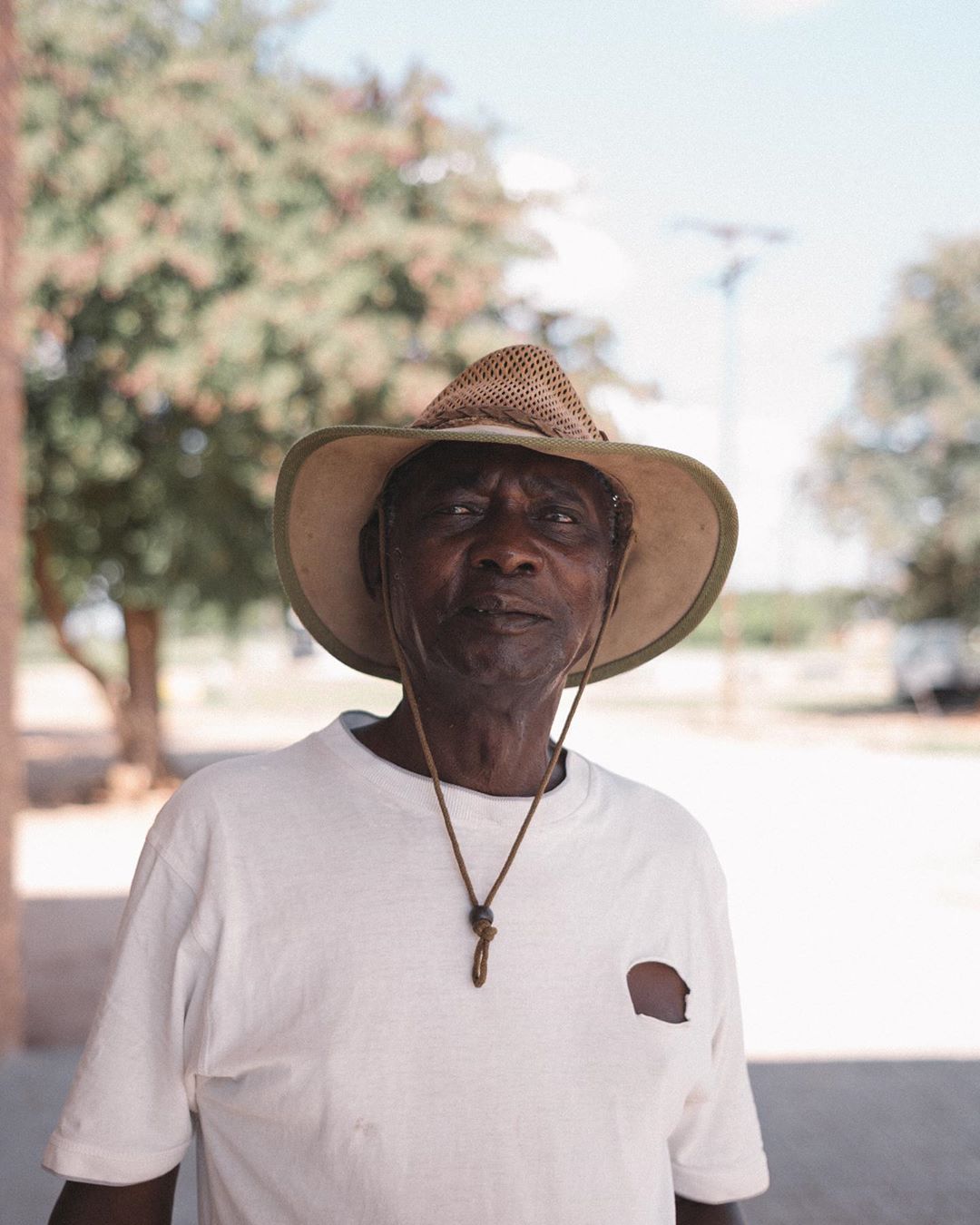Older African-American man wearing a straw hat and white t-shirt stands outdoors. Portrait captures his face and upper body in soft light.
