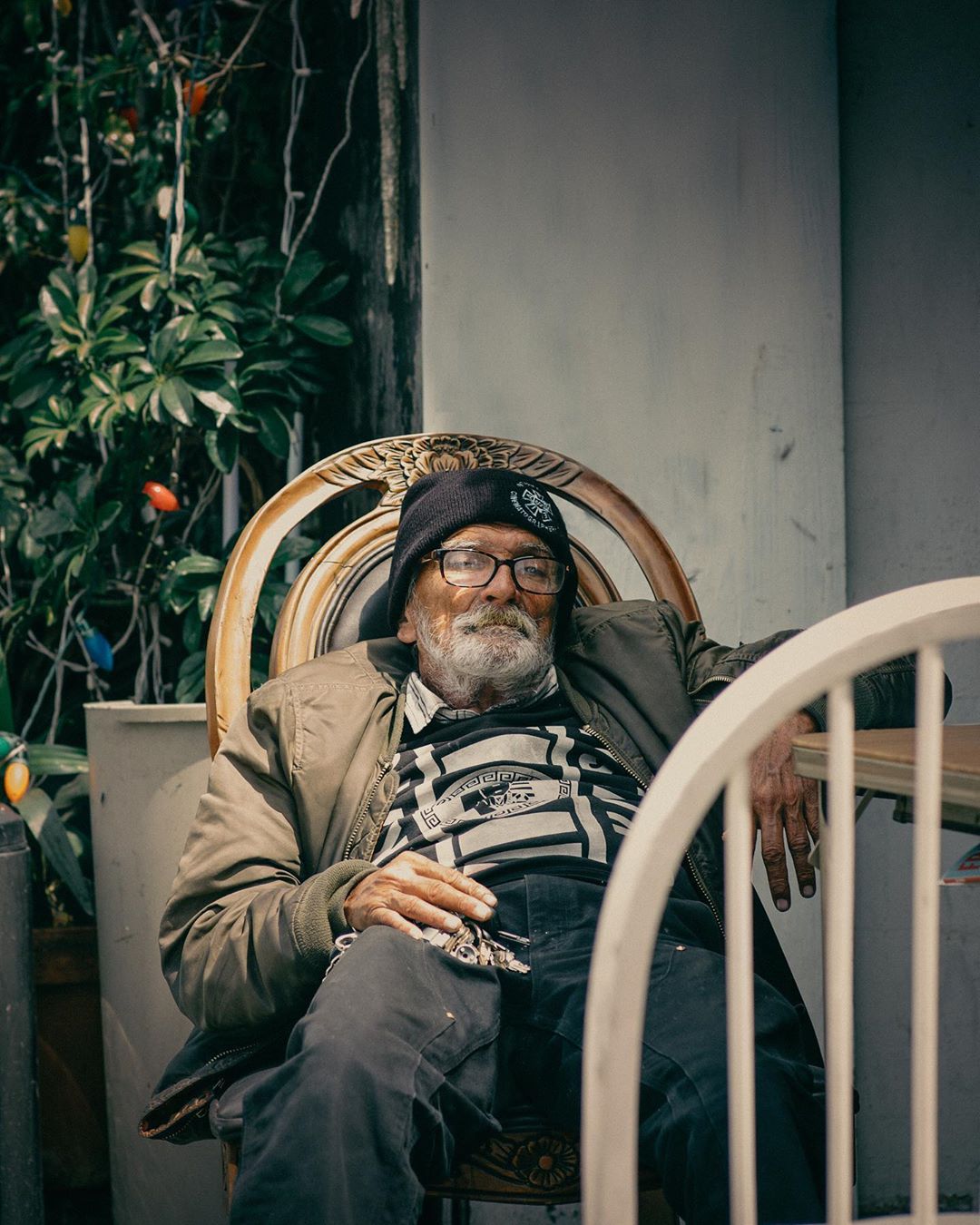 Elderly man with beard, wearing glasses and beanie, relaxes in an ornate chair on a porch, surrounded by greenery and a weathered wall.