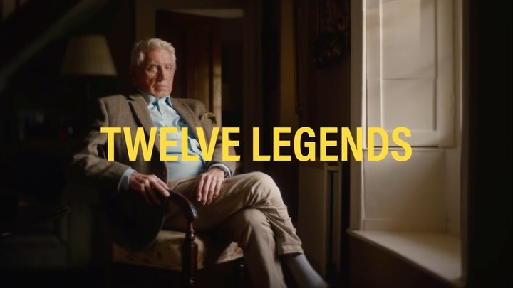 Older Caucasian man in a tweed jacket sits in a chair indoors next to a window with the text 'Twelve Legends' superimposed.