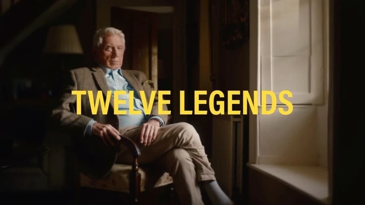 Older Caucasian man in a tweed jacket sits in a chair indoors next to a window with the text 'Twelve Legends' superimposed.