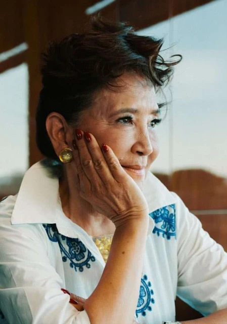 Mature woman in elegant white and blue patterned shirt with hand near face, gold earrings, and red nails gazes out of frame.
