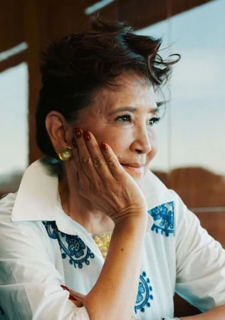 Elderly woman with short dark hair resting her hand on her cheek, wearing a white patterned blouse.