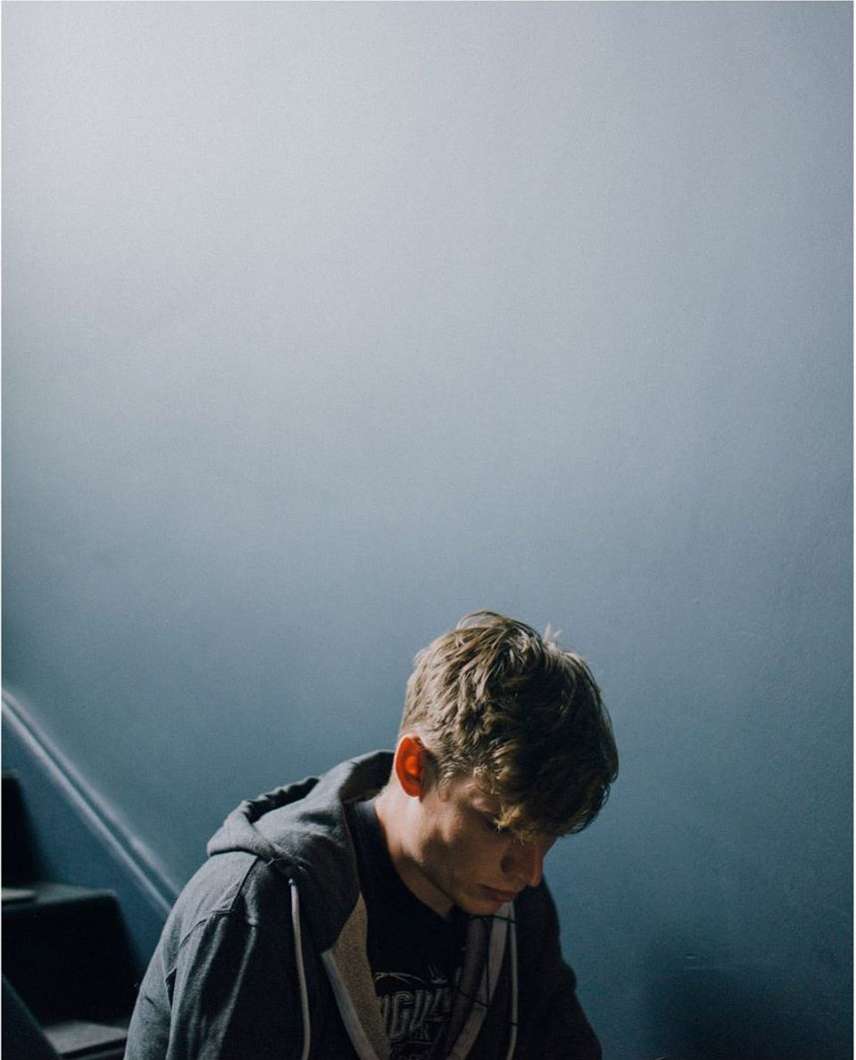 Young adult man with blonde hair in a hooded sweatshirt sits on stairs indoors with a light blue wall and dark staircase in the background.