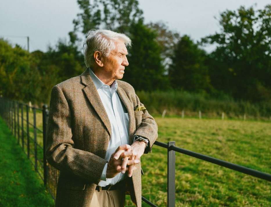Elderly man in tweed jacket, standing outdoors, looking away. He is near a fence, with a grassy field and trees in the background.