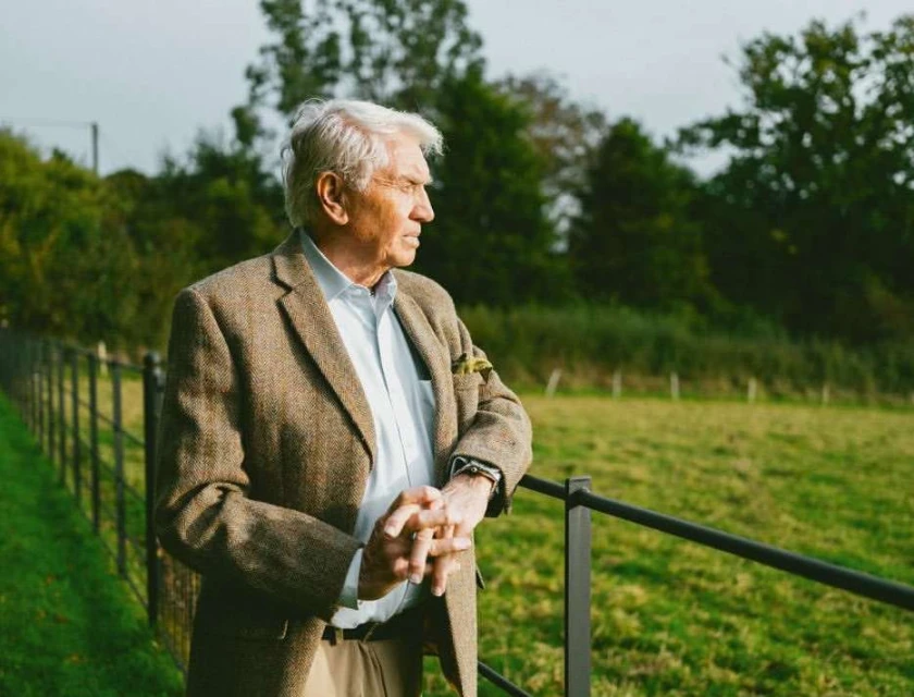 Elderly man in tweed jacket, standing outdoors, looking away. He is near a fence, with a grassy field and trees in the background.