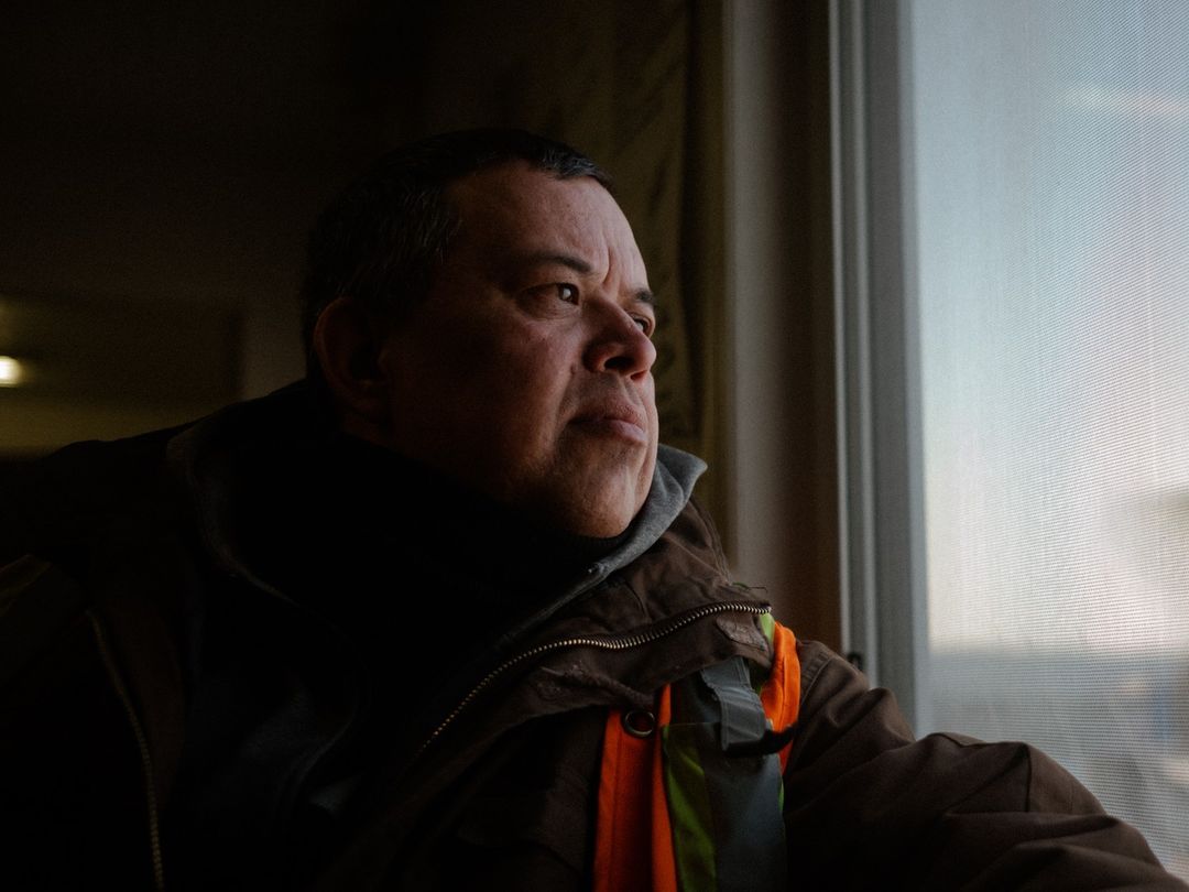 Indoor portrait of an adult male wearing a brown jacket and safety vest, looking out a window with natural light illuminating his face.