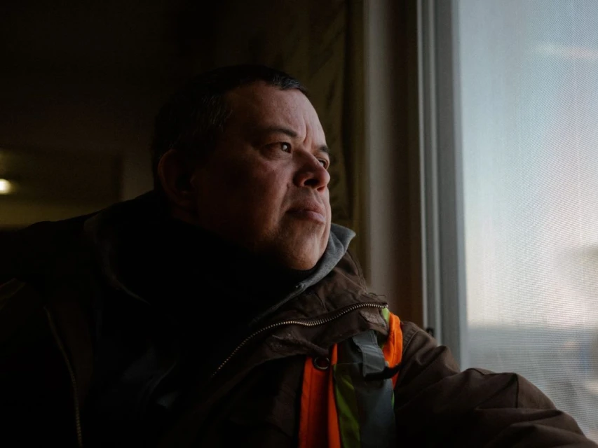Indoor portrait of an adult male wearing a brown jacket and safety vest, looking out a window with natural light illuminating his face.