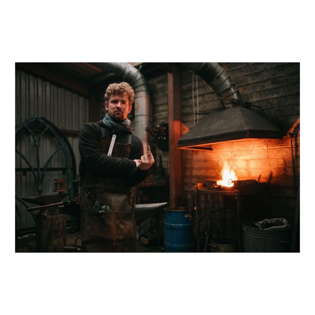 A man in a blacksmith shop stands in front of a furnace with a metal rod being heated, wearing a leather apron, inside a metal and wood building.