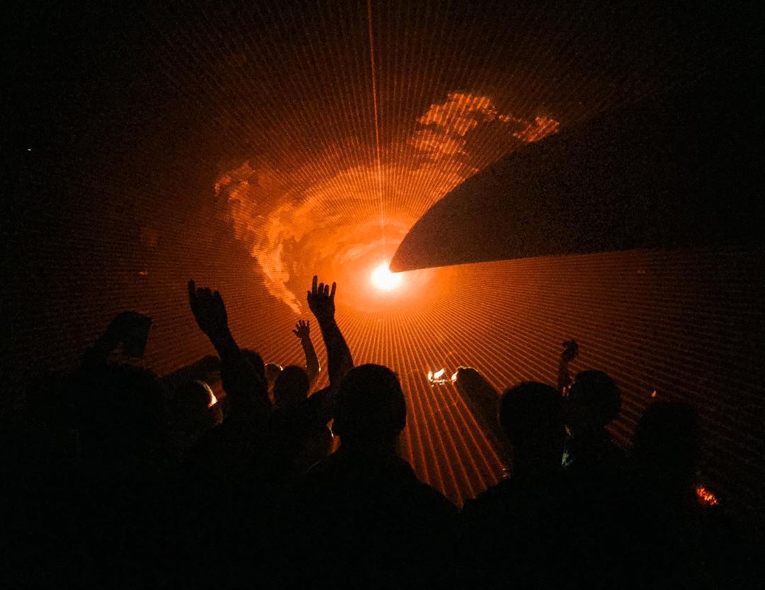 Silhouetted crowd at a concert illuminated by bright orange laser lights and stage lighting creating an energetic atmosphere.
