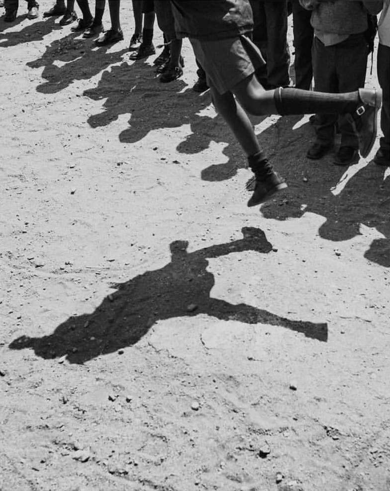 A group of young people, mostly legs and shadows, are walking or running across a sandy surface in a black and white photo.