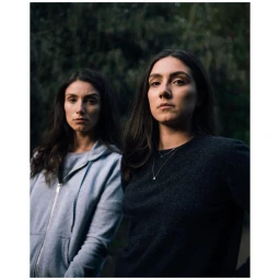 Two women with long dark hair standing outdoors with a dark foliage background, photographed by Ben Perry in London.