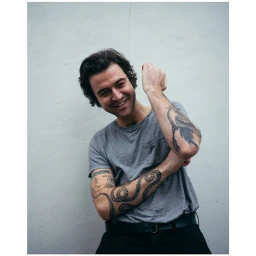 Portrait of a smiling man with heavily tattooed arms wearing a grey t-shirt against a plain white wall