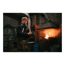 A blacksmith wearing a leather apron and dark hoodie stands in a workshop with a lit forge in the background, photographed by Ben Perry.