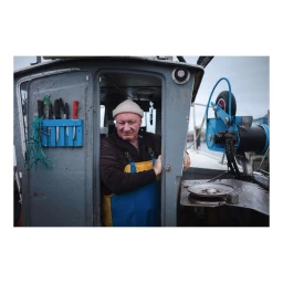 Fisherman in white beanie and blue and yellow overalls standing inside the cabin of a fishing boat by Ben Perry