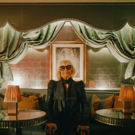 An elderly woman wearing dark sunglasses and a black leather jacket sitting on a sofa between two small lamps.