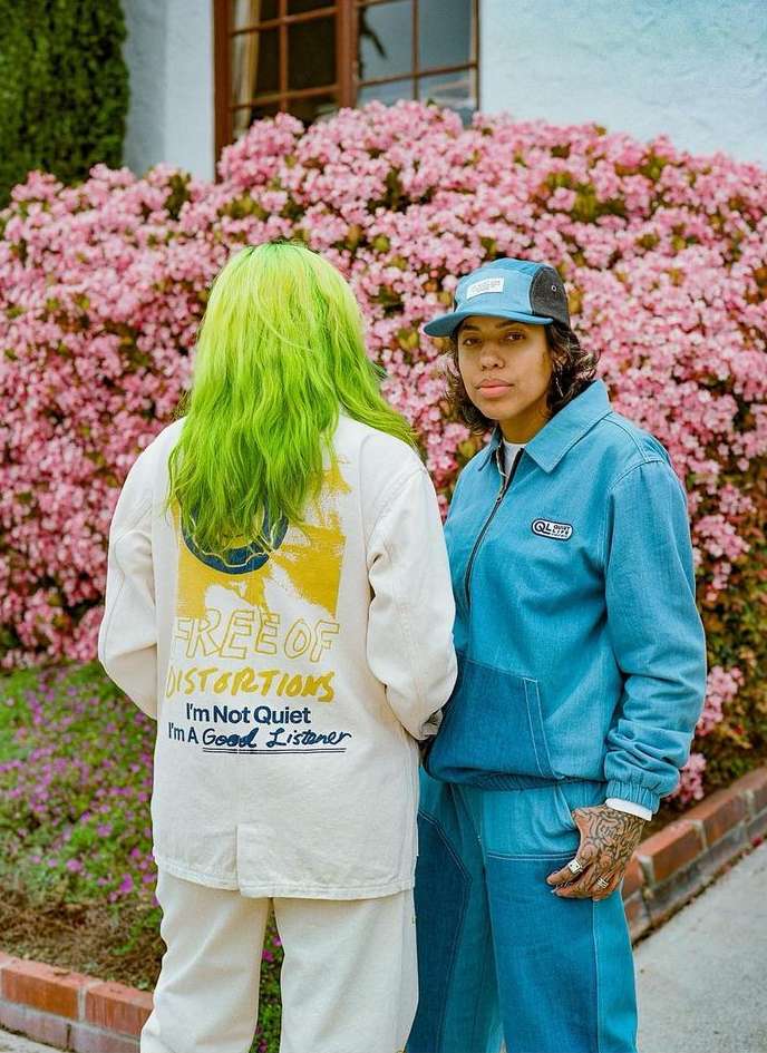 Two people stand outdoors next to a blooming azalea bush. One person wears green hair, the other wears a blue baseball cap.