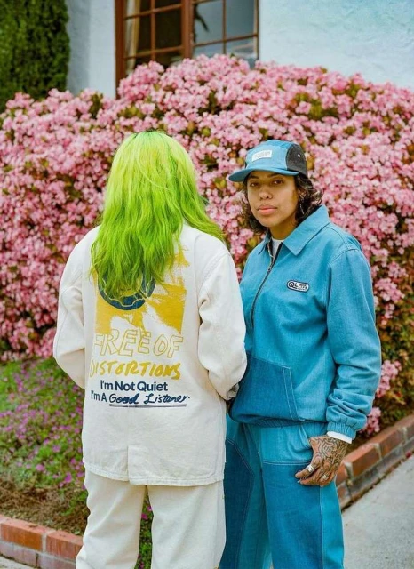 Two people stand outdoors next to a blooming azalea bush. One person wears green hair, the other wears a blue baseball cap.