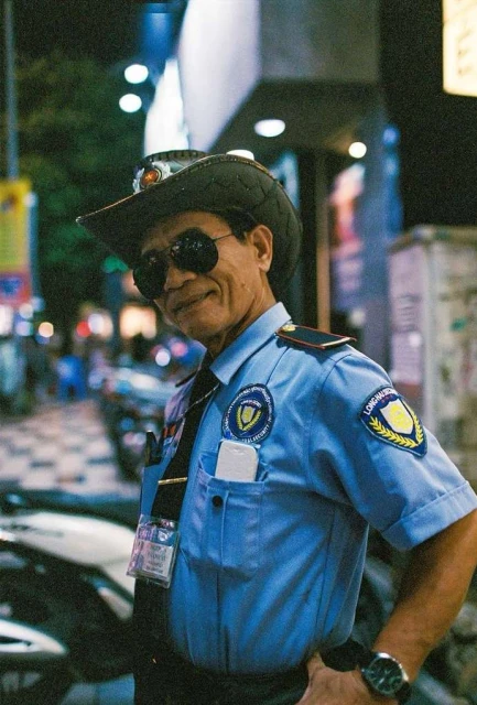 An older man poses at night in a security guard uniform and cowboy hat, with a dark tie, sunglasses, and watch.