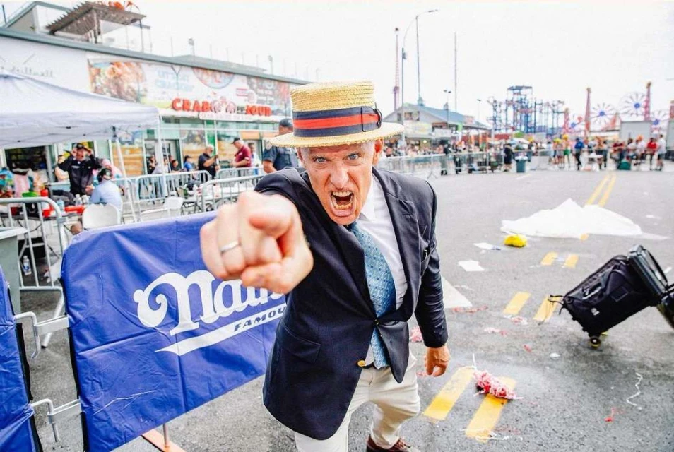 Man in straw hat and blazer points at the camera outdoors on street with banner and distant crowd and food vendors in Coney Island.